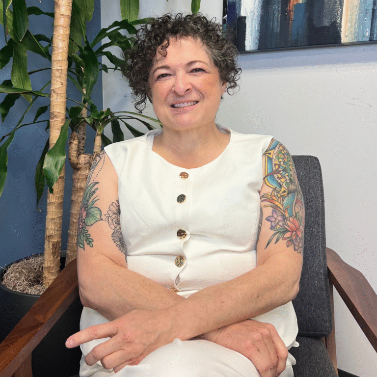 Woman seated in a chair smiling with short, dark curly hair and a white blouse.